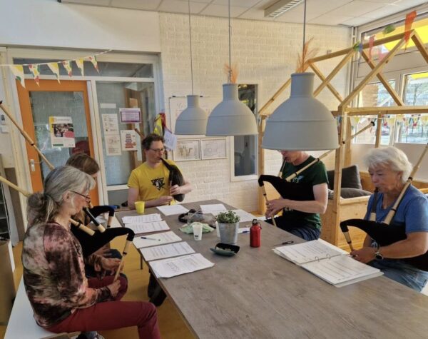 Vijf mensen zitten rond een tafel in een klaslokaal. Ze spelen allemaal doedelzak met de docent Menno aan het hoofd van de tafel. Op de tafel ligt bladmuziek.